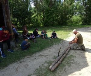 Dzieci znajdują się na świeżym powietrzu. Prowadzący pokazuje jak wykonać pułapkę na zwierzę.