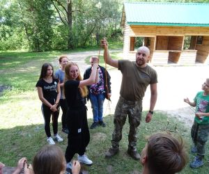 Dzieci znajdują się na świeżym powietrzu. Prowadzący pokazuje jak wykonać pułapkę na zwierzę.