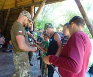 Dzieci znajdują się na świeżym powietrzu. Prowadzący pokazuje jak wykonać pułapkę na zwierzę.
