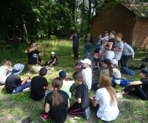 Dzieci siedzą w okręgu na świeżym powietrzu na polanie. Prowadzący zajęcia znajduje tłumaczy coś dzieciom.