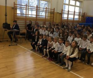 Uczniowie siedzą na ławkach przed występującymi uczniami