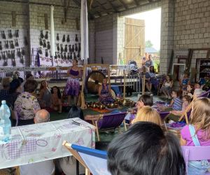 Zdjęcie przedstawia publiczność na koncercie, który odbywa się w stodole, a na środku panią, która gra na misach.