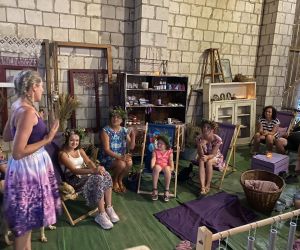 Zdjęcie przedstawia publiczność na koncercie, który odbywa się w stodole, a z boku stoi kobieta, która przemawia do publiczności.