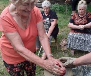Zdjęcie przedstawia osobę znajdująca się na świeżym powietrzu, która lepi garnek z gliny. Obok pomaga instruktor.