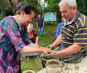 Zdjęcie przedstawia osobę znajdująca się na świeżym powietrzu, która lepi garnek z gliny. Obok pomaga instruktor.