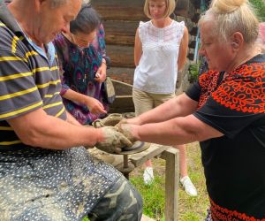 Zdjęcie przedstawia osobę znajdująca się na świeżym powietrzu, która lepi garnek z gliny. Obok pomaga instruktor.