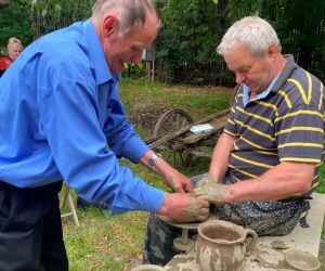 Zdjęcie przedstawia osobę znajdująca się na świeżym powietrzu, która lepi garnek z gliny. Obok pomaga instruktor.