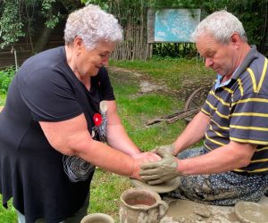 Zdjęcie przedstawia osobę znajdująca się na świeżym powietrzu, która lepi garnek z gliny. Obok pomaga instruktor.