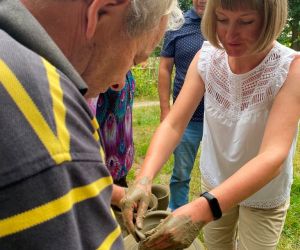 Zdjęcie przedstawia osobę znajdująca się na świeżym powietrzu, która lepi garnek z gliny. Obok pomaga instruktor.