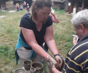 Zdjęcie przedstawia osobę znajdująca się na świeżym powietrzu, która lepi garnek z gliny. Obok pomaga instruktor.