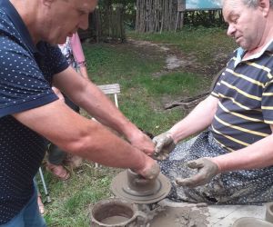 Zdjęcie przedstawia osobę znajdująca się na świeżym powietrzu, która lepi garnek z gliny. Obok pomaga instruktor.