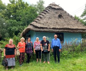 Zdjęcie przedstawia grupę ludzi na świeżym powietrzu stojących przed chata z drewna.