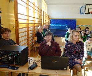 Uczniowie z Samorządu Uczniowskiego wraz z opiekunem siedzą przy sprzęcie muzycznym nagłaśniającym, w tle nauczyciele i uczniowie