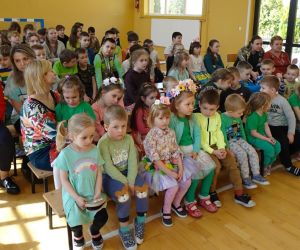 Najmłodsze dzieci w wiosennych strojach siedzą na ławce w pierwszym rzędzie, w sali gimnastycznej