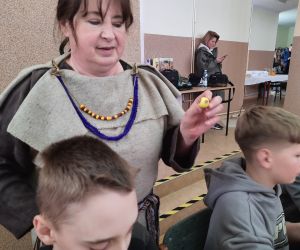 Kobieta w gockim stroju pomaga dzieciom w wykonaniu paciorków z modeliny