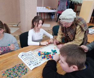 Kobieta w gockim stroju pomaga uczennicy przy wykonywaniu paciorka z modeliny
