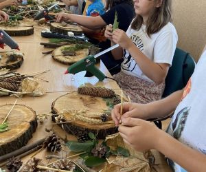 Zdjęcie przedstawia grupę dzieci wewnątrz budynku. Dzieci wykonują pracę plastyczną - dekoracyjny plaster drewna.