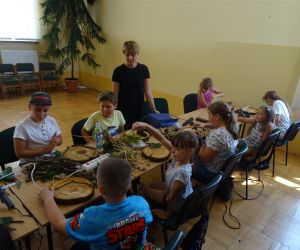 Zdjęcie przedstawia grupę dzieci wewnątrz budynku oraz jedna osobę dorosłą. Dzieci wykonują pracę plastyczną - ozdobny plaster drewna.
