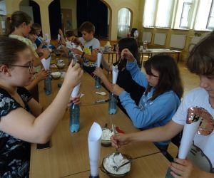 Zdjęcie przedstawia grupę dzieci wewnątrz budynku oraz osobę dorosłą. Dzieci wykonują 