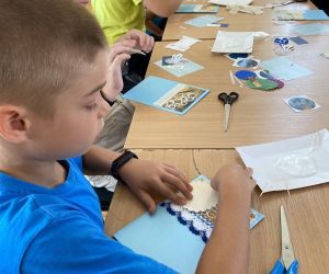 Zdjęcie przedstawia grupę dzieci wewnątrz budynku. Dzieci wykonują kartkę z wakacji metoda scrapbookingu.