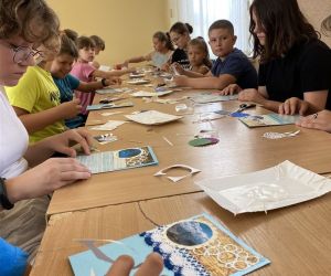 Zdjęcie przedstawia grupę dzieci wewnątrz budynku. Dzieci wykonują kartkę z wakacji metoda scrapbookingu.