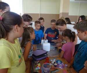 Zdjęcie przedstawia grupę dzieci wewnątrz budynku oraz osobę dorosłą. Dzieci wykonują 