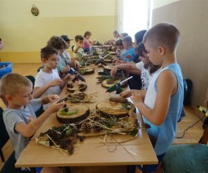 Zdjęcie przedstawia grupę dzieci wewnątrz budynku. Dzieci wykonują prace plastyczną - dekoracyjny plaster drewna.