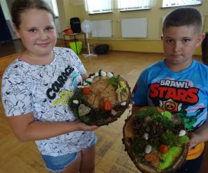 Zdjęcie przedstawia dwoje dzieci wewnątrz budynku. Dzieci prezentują swoje prace plastyczne - ozdobiony plaster drewna.
