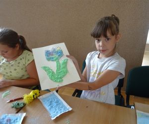 Zdjęcie przedstawia dzieci wewnątrz budynku. Dziecko prezentuje prace plastyczną - malowanie solą.