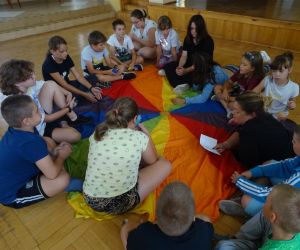 Zdjęcie przedstawia grupę dzieci wewnątrz budynku. Dzieci bawią się chustą animacyjną.