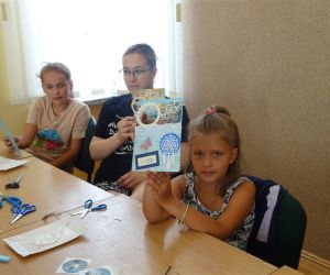 Zdjęcie przedstawia grupę dzieci wewnątrz budynku. Dzieci prezentują kartki z wakacji, które wykonały samodzielnie podczas warsztatów.