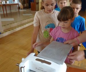 Zdjęcie przedstawia grupę dzieci wewnątrz budynku. Dzieci obsługują maszynę tnąco-wytłaczającą służącą do scrapbookingu.