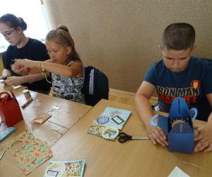 Dzieci prezentują pudełka na skarby, które wykonały samodzielnie podczas warsztatów. Dzieci znajdują się wewnątrz budynku.