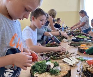 Zdjęcie przedstawia grupę dzieci wewnątrz budynku. Dzieci wykonują prace plastyczną - dekoracyjny plaster drewna.