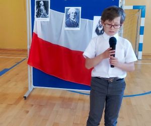 Uczeń klasy czwartej recytuje przy mikrofonie na tle tablicy dekoracyjnej