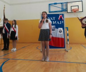 Uczennica klasy czwartej i uczeń klasy siódmej przy mikrofonach, w tle poczet sztandarowy