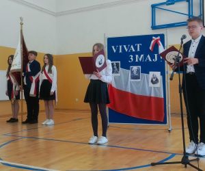 Uczennica klasy piątej i uczeń klasy siódmej recytują przy mikrofonach, w tle poczet sztandarowy