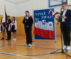 Uczennica klasy czwartej i uczeń klasy siódmej przy mikrofonach, w tle poczet sztandarowy