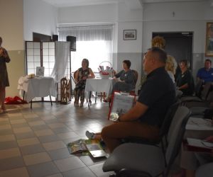 Na zdjęciu widoczna jest grupa osób siedząca na krzesłach i jedna osoba mówiąca do mikrofonu.