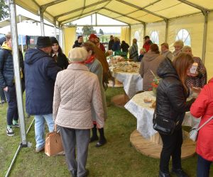 Na zdjęciu znajdują się osoby na świeżym powietrzu będące pod namiotem przy stoisku z ciastem.