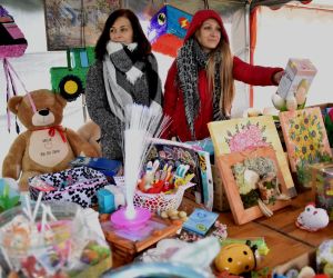 Na zdjęciu znajdują się dwie kobiety, na powietrzu pod namiotem, stojące przy stoisku rękodziełem, maskotkami i innymi przedmiotami przekazanymi na kiermasz charytatywny.