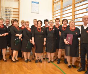 Zespół śpiewaczy z Instruktorem stoją przed scena wewnątrz budynku.